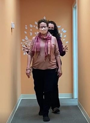 Two women walking in a hallway as part of a training exercise with prism glasses, with one person guiding the other from behind.