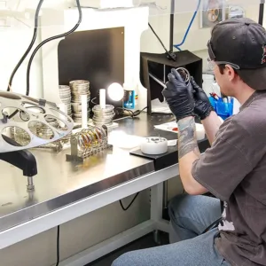 Lab technician seated at a workbench, inspecting a lens with various optical tools and equipment around.