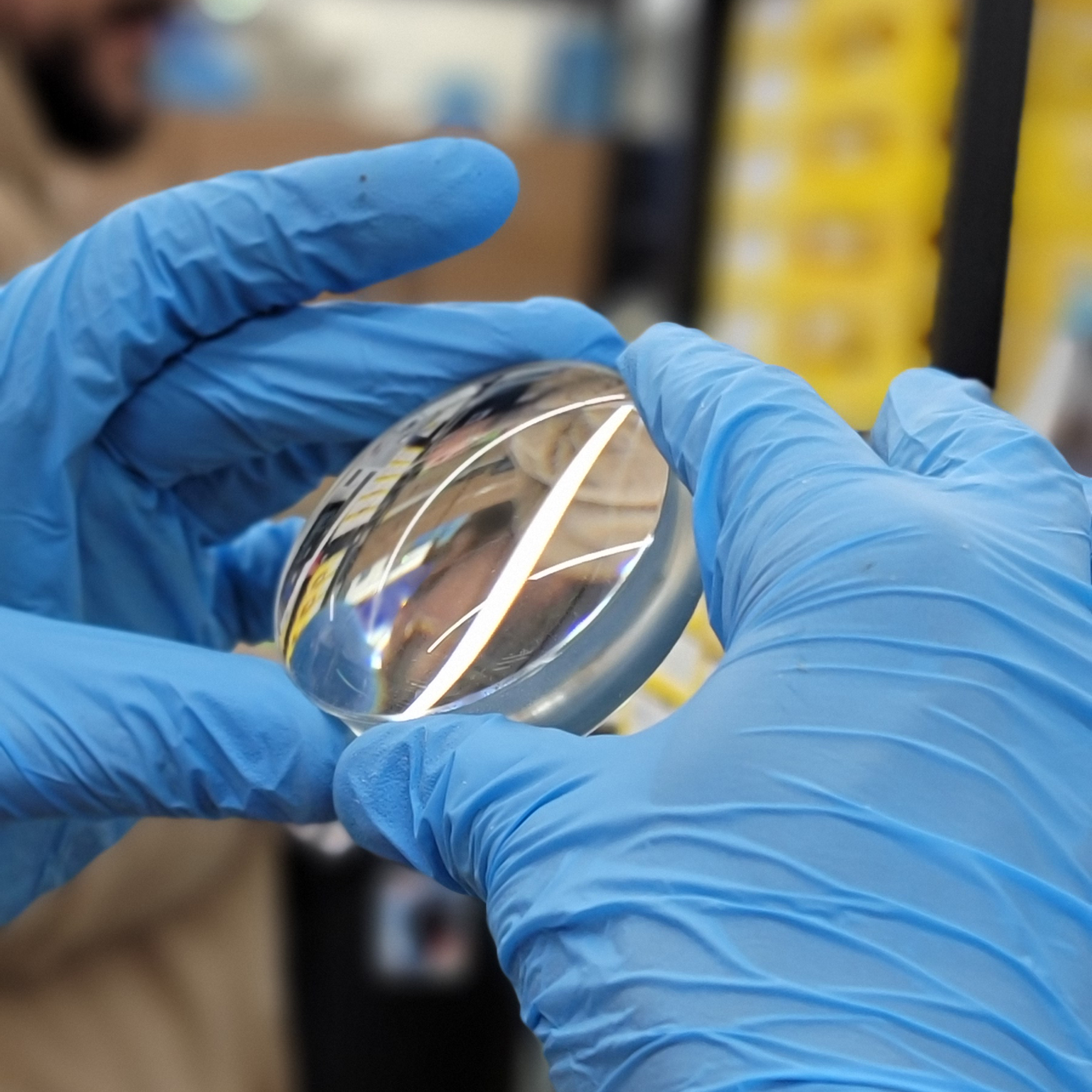 Gloved hands holding a semi-finished lens, showing its reflective surface under bright lighting in a lens manufacturing setting