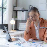 Woman in an office environment massaging her eyes in fatigue, with glasses in hand, representing eye strain from screen use.