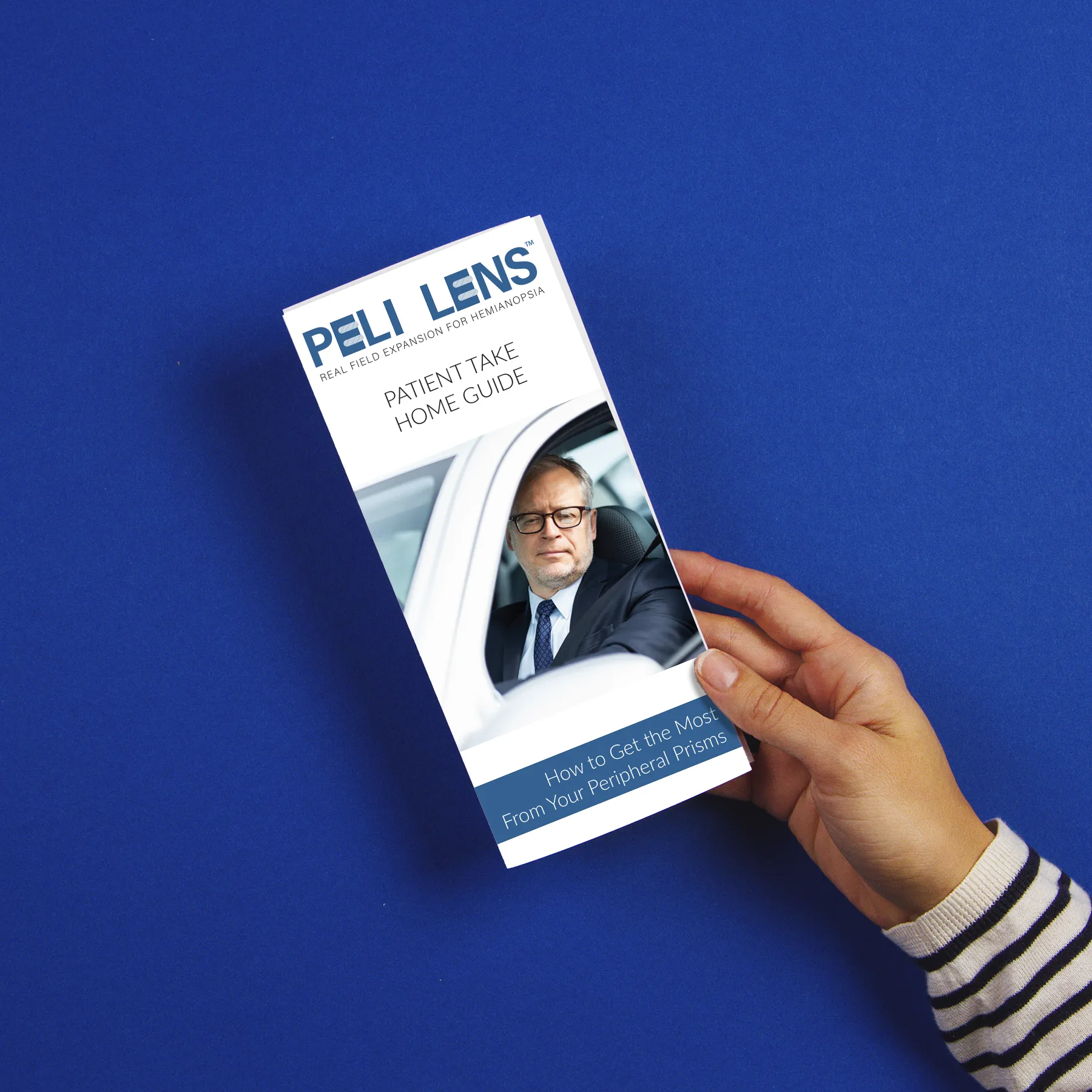 A person holds a brochure titled "Peli Lens: Patient Take Home Guide" with a man in a suit pictured on the cover, against a blue background.