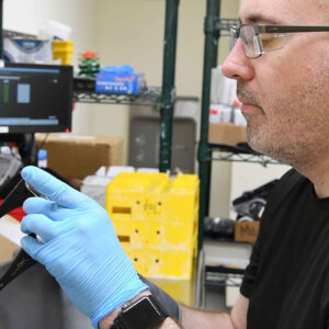 Bryan examines a lens in the Quality Control area of Chadwick Optical, wearing blue gloves and focused on the inspection, with lab equipment and storage boxes in the background.