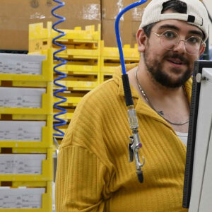 Matt, standing near stacked trays and a monitor in Chadwick Optical’s lab, smiles toward the camera, with a blue air hose coiled overhead, part of the equipment setup in the workspace.