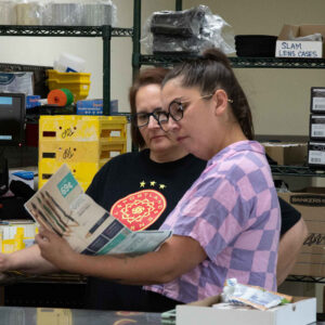 Jenn and Jordan examine a catalog together in Chadwick Optical’s lab area, surrounded by organized supplies and equipment, likely discussing product options or inventory.