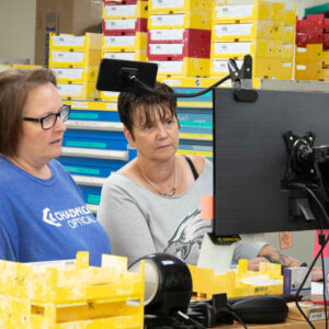 Jenn (left) and Susan (right) examine a computer screen together in Chadwick Optical’s lab, surrounded by trays of organized lenses, discussing work-related details.