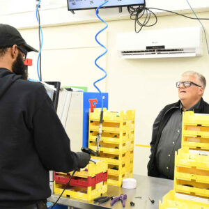 Ken and Ralf at Chadwick Optical discuss tasks at a workstation surrounded by yellow trays and tools.