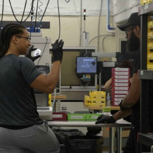 Esmerlin (left) and a colleague engage in conversation at Chadwick Optical’s lab, surrounded by equipment and organized trays used for processing lenses.