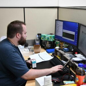 Alex at Chadwick Optical seated at his desk, working on a computer with a dual monitor setup, surrounded by office items and various personal desk decorations.