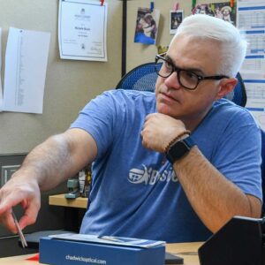 Tom seated at his desk at Chadwick Optical, pointing to a small item, with office supplies and documents around him.