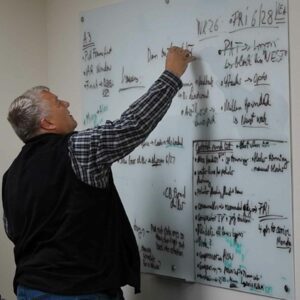 Man in a plaid shirt and black vest writes on a large whiteboard covered with handwritten notes and diagrams, appearing focused on the task.