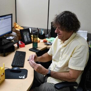 Tom at Chadwick Optical seated at his desk, working with small parts in his hands, with his computer and office items arranged on the desk around him.