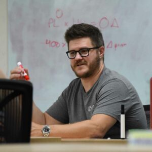 Charlie holding a red marker, looking engaged during a meeting at Chadwick Optical, with equations and notes on the whiteboard behind him.