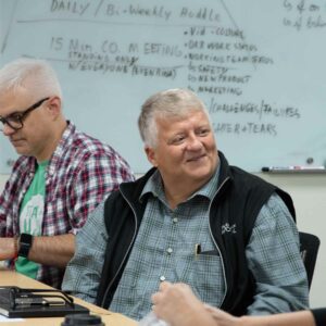 Ralf seated at a meeting table at Chadwick Optical, smiling with a whiteboard full of notes and topics listed in the background.