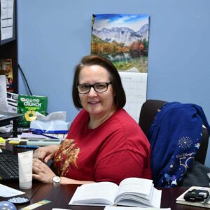 Jenn sitting at her desk with open books and documents, a calendar on the wall, working at Chadwick Optical.