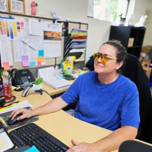 Woman working at a desk with yellow tinted fit-over glasses, using a computer in an office setting.