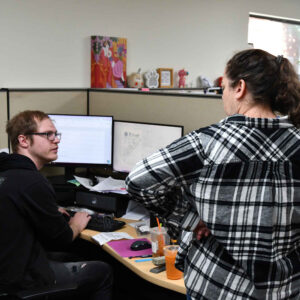 John and Amanda at Chadwick Optical, engaged in discussion at John's desk workstation, with dual monitors displaying work documents.