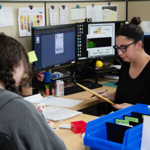 Faith and Jordan working together at a desk, with a computer screen showing shipping labels and workspace materials surrounding them.