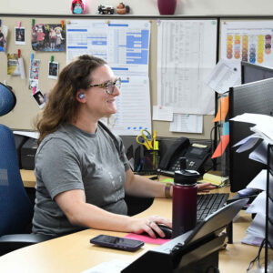 Michelle at Chadwick Optical, working at her desk and focused on her computer screen, with a background of pinned notes and schedules on the cubicle wall.