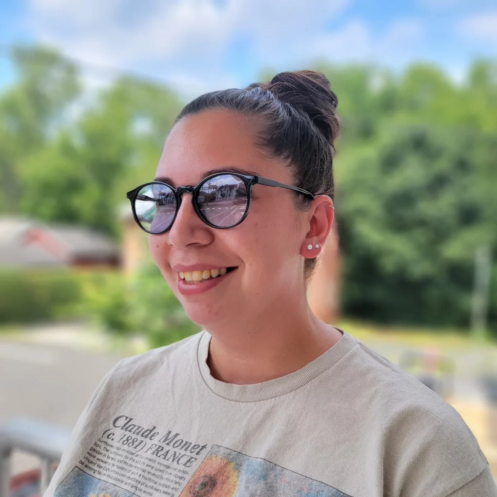 Smiling woman wearing round, tinted glasses with a casual Claude Monet t-shirt, standing outdoors with greenery in the background.