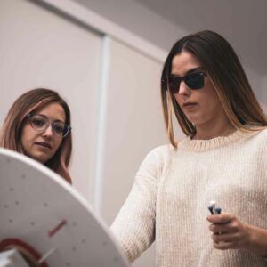 Two women engaging in a vision training exercise, one wearing tinted glasses and holding a pointer tool, with focused expressions.