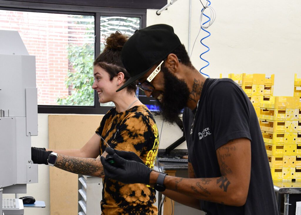 Two employees smiling and focused on their tasks in a workshop, with one holding a tool, surrounded by yellow bins and equipment.