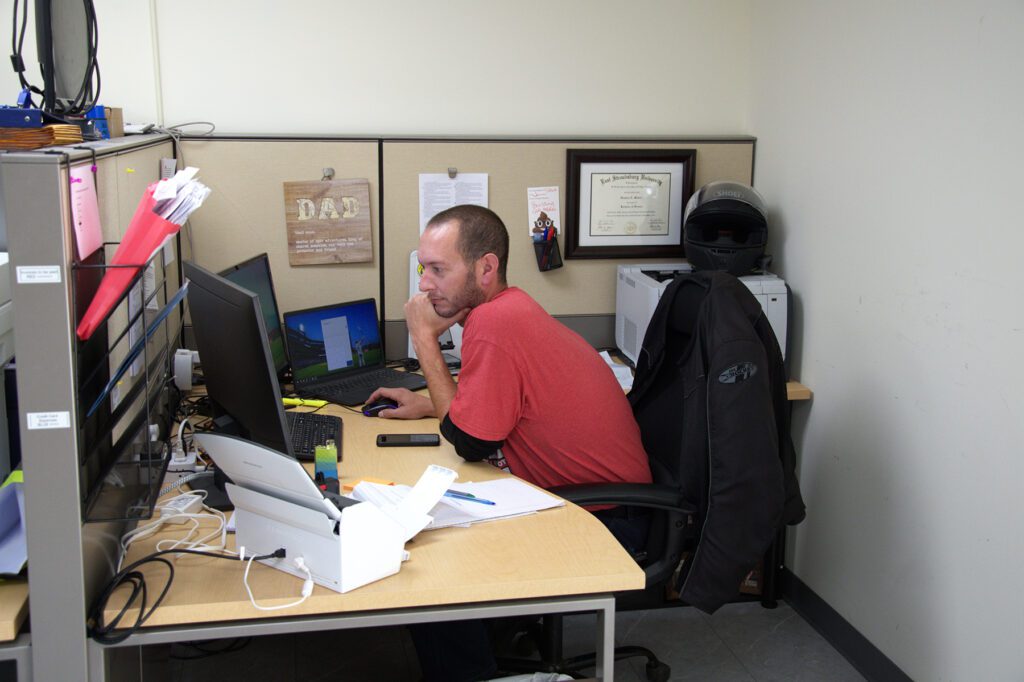 Man in a red shirt focused on his computer at a cubicle desk, with a motorcycle helmet and framed diploma on the wall behind him.