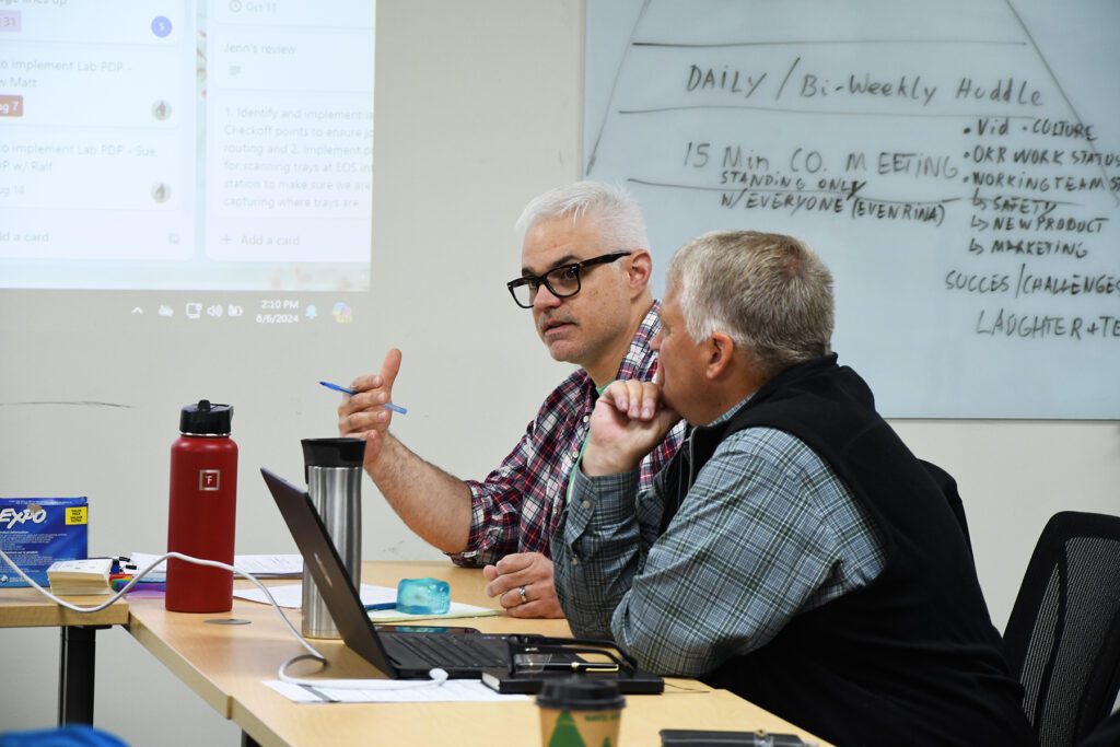 Two men engaged in a discussion during a meeting at a conference table, with notes visible on a whiteboard in the background.