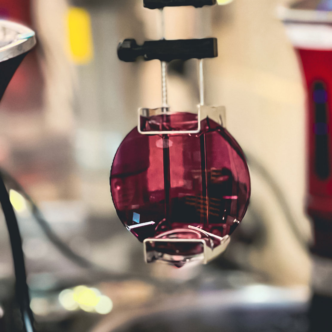 Red-tinted lens held in a clamp during a tinting process, with a blurred laboratory background.