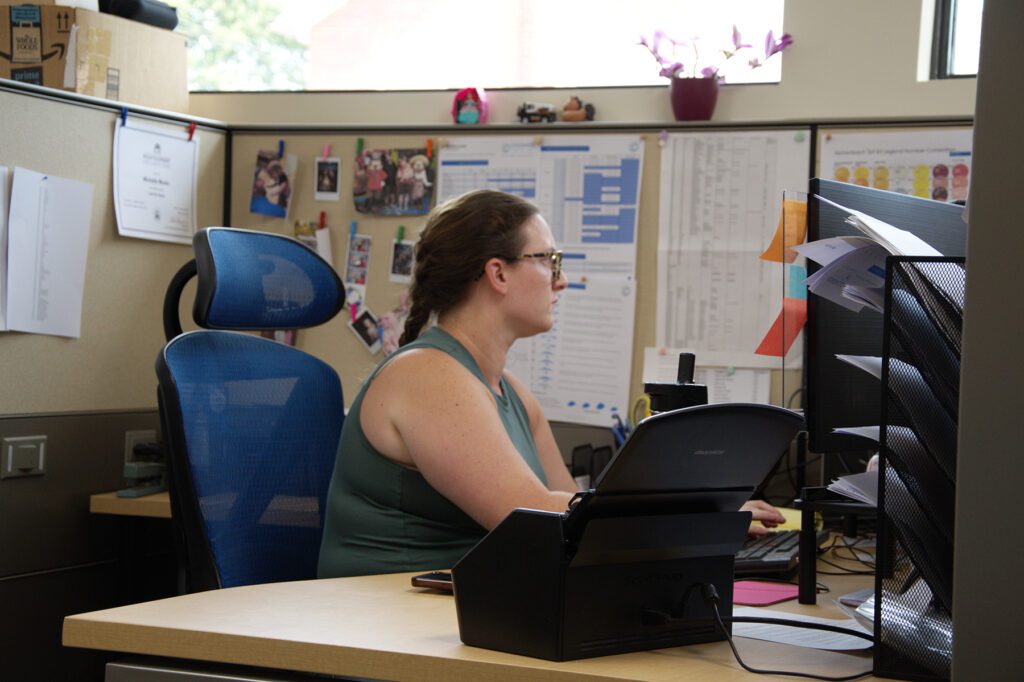 Woman in a cubicle working on a computer, surrounded by office materials, family photos, and charts on the wall, at Chadwick Optical.