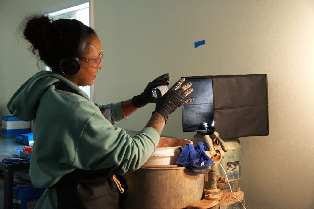 Woman wearing safety goggles and gloves working on a lens casting process under focused lighting, with hands positioned over casting equipment at Chadwick Optical.