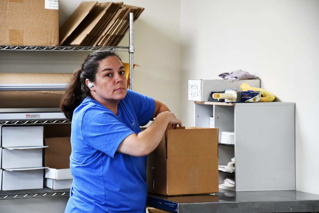 Woman in a blue shirt, wearing an earbud, packing a cardboard box in a workspace with shelves and packing materials.