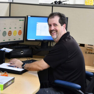 Ed, Shipping Manager, smiling at his desk with dual monitors displaying shipping information.