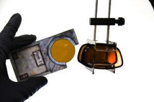 Close-up of a lens being tested with a yellow-tinted filter in a metal holder, against a plain background.