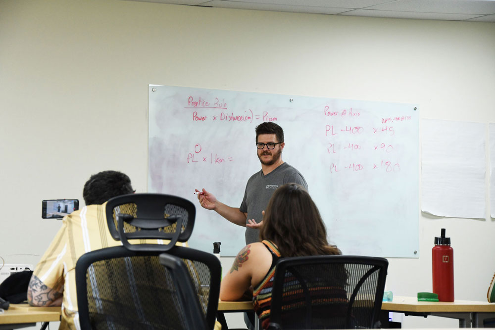 Charlie leads a training session at Chadwick Optical, explaining equations on a whiteboard to a group of engaged employees.