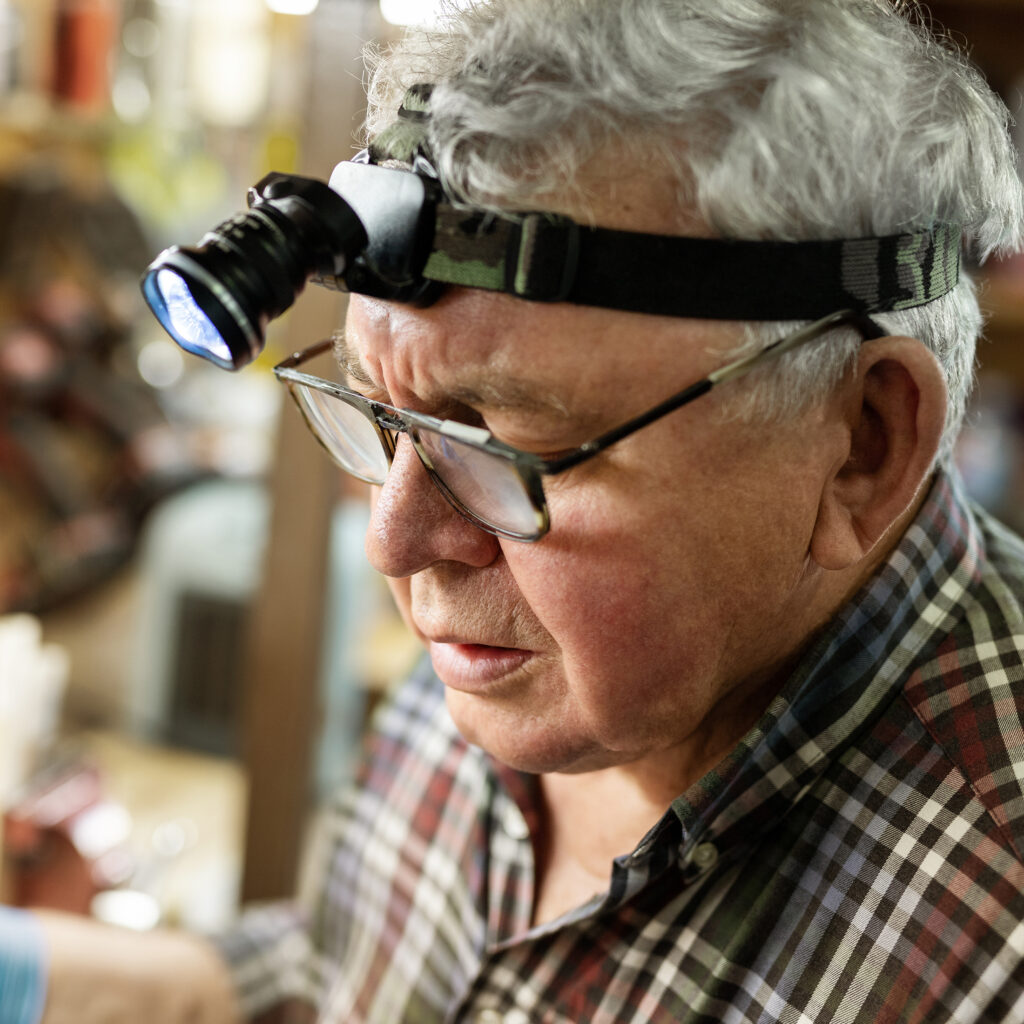 Senior,Craftsman,Portrait,With,Headlamp,Led,Torch,Working,In,Workhop Senior craftsman portrait with headlamp led torch working in workhop with tools and equipment on background. Old mature gray handyman tutor with eyeglasses. Experinced person making diy hobby craft
