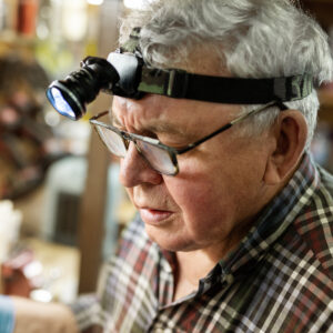 Senior craftsman portrait with headlamp led torch working in workhop with tools and equipment on background. Old mature gray handyman tutor with eyeglasses. Experinced person making diy hobby craft