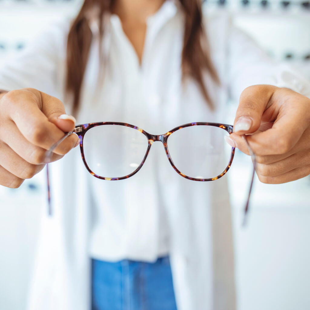 Optician showing and suggesting eyeglasses in optical shop. Cheerful female ophthalmologist is working with patient. Optician giving new glasses to customer for testing and trying.