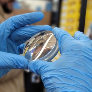 Technician in blue nitrile gloves holding a thick round high-plus lens blank up to the light for inspection.