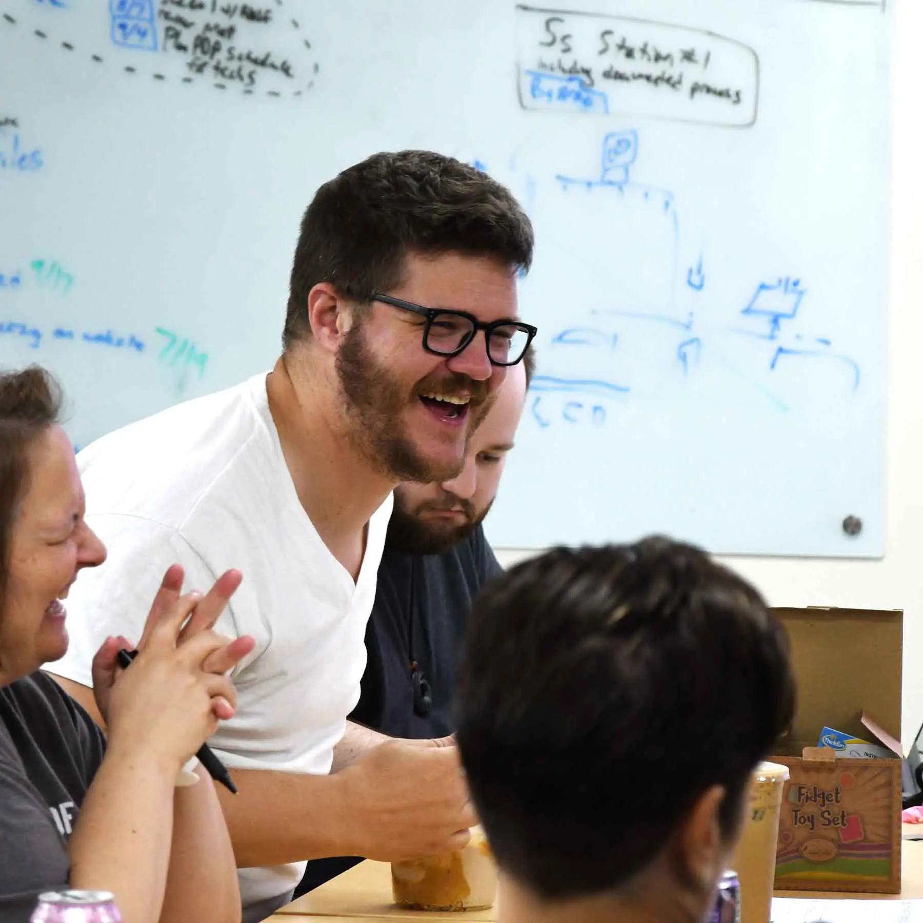 Team members laughing during a meeting in front of a whiteboard covered in handwritten notes and diagrams.