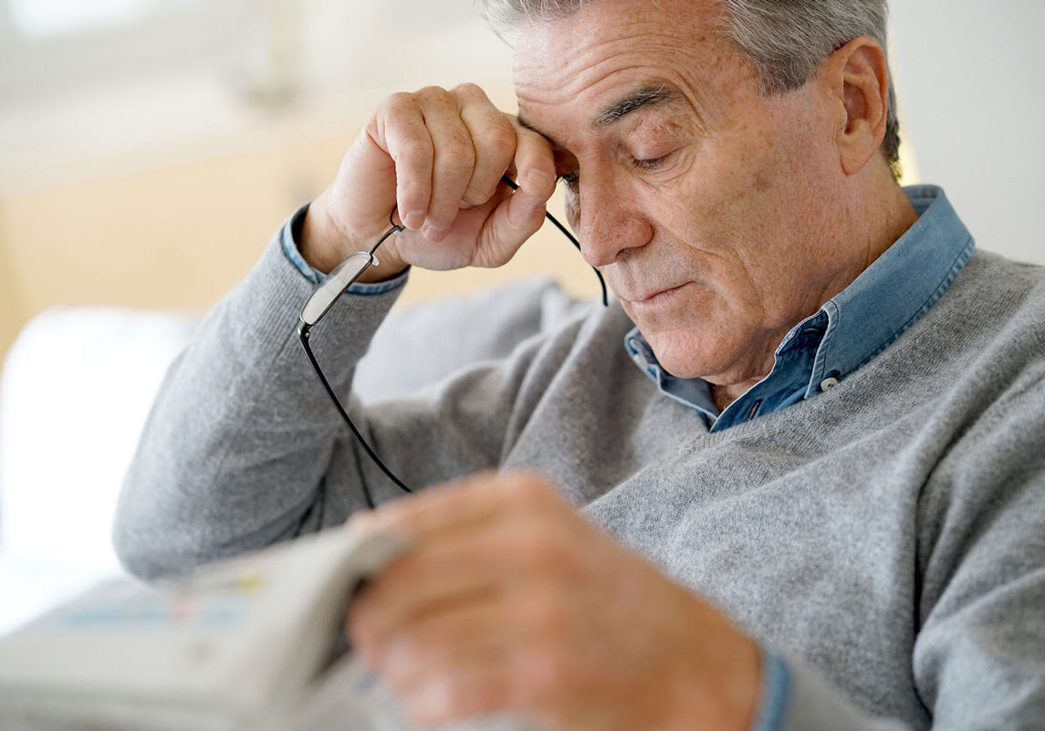 Binocular_Vision_Dysfunction4_Chadwick_Optical Older man removing his glasses while squinting, appearing fatigued or strained, with a newspaper in his hand.
