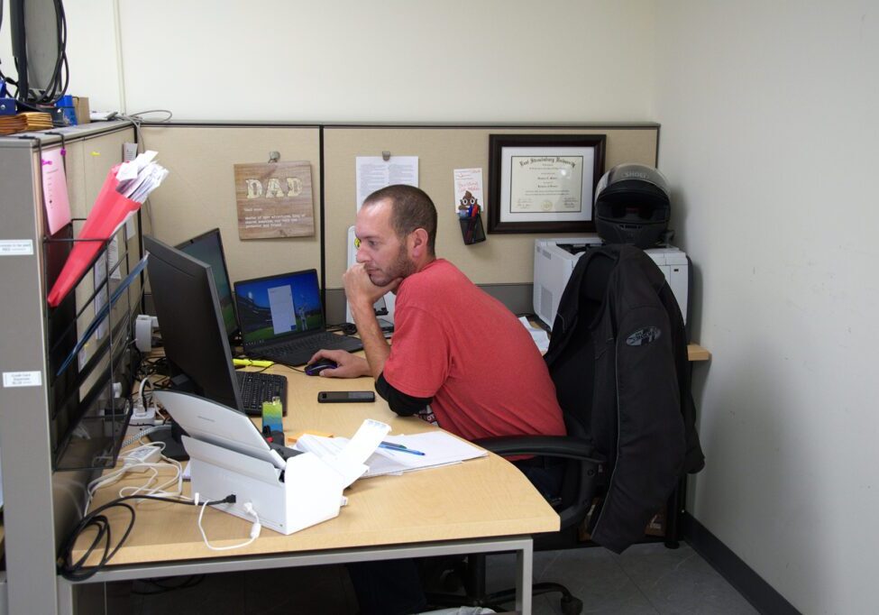 Man in a red shirt focused on his computer at a cubicle desk, with a motorcycle helmet and framed diploma on the wall behind him.
