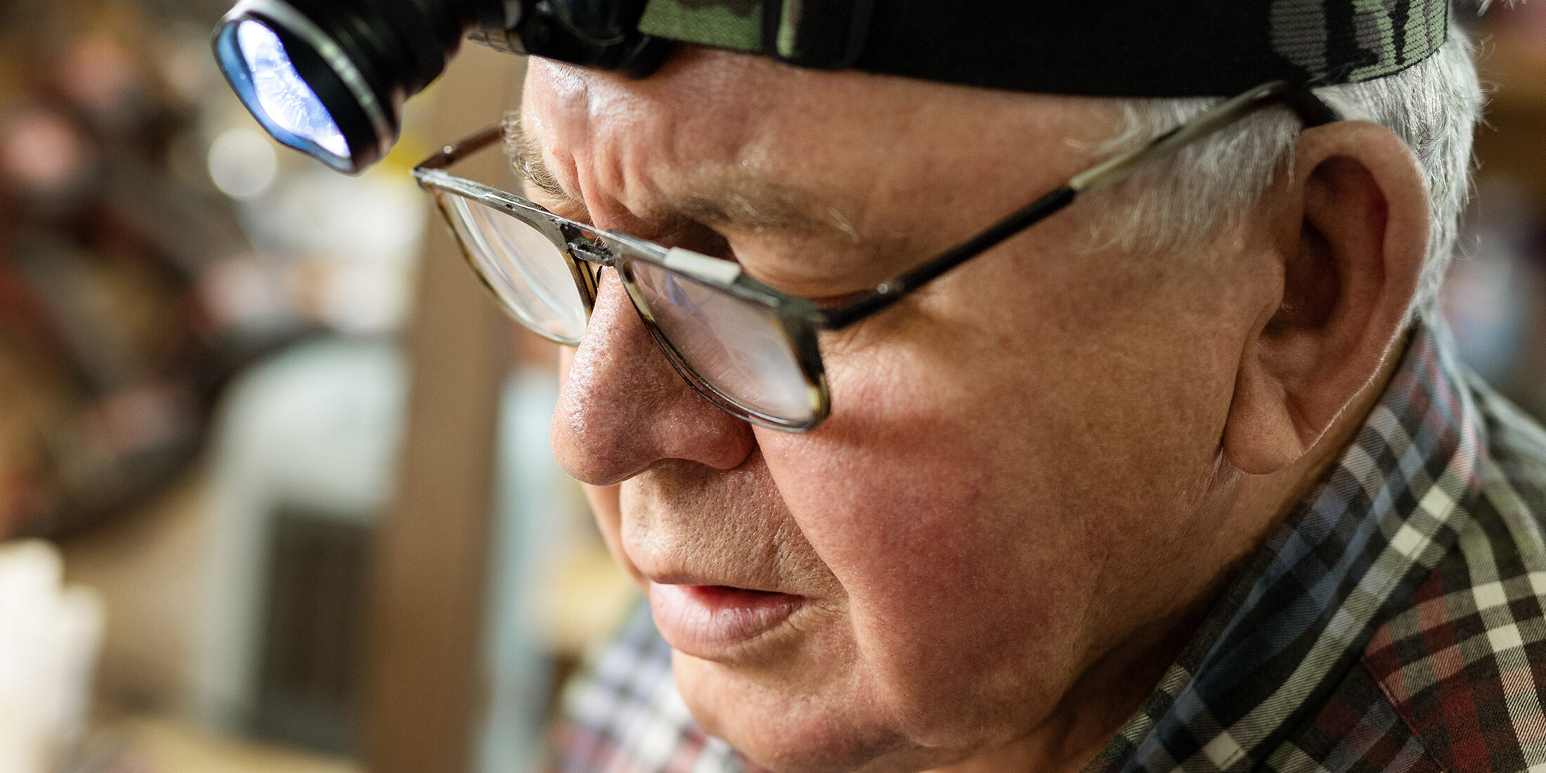 Senior craftsman portrait with headlamp led torch working in workhop with tools and equipment on background. Old mature gray handyman tutor with eyeglasses. Experinced person making diy hobby craft