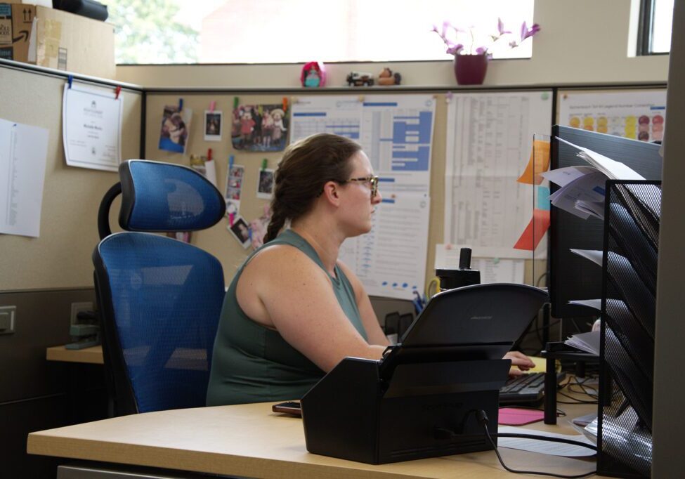 Woman in a cubicle working on a computer, surrounded by office materials, family photos, and charts on the wall, at Chadwick Optical.