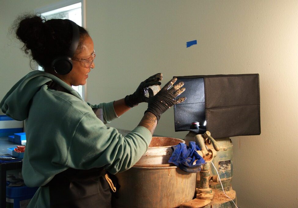 Woman wearing safety goggles and gloves working on a lens casting process under focused lighting, with hands positioned over casting equipment at Chadwick Optical.
