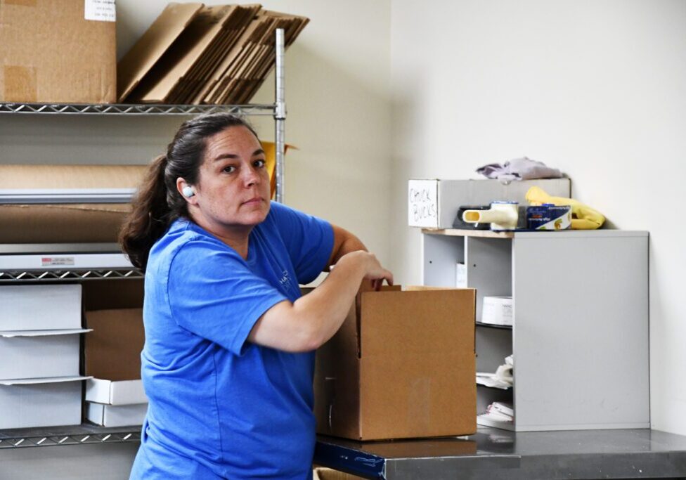 Woman in a blue shirt, wearing an earbud, packing a cardboard box in a workspace with shelves and packing materials.